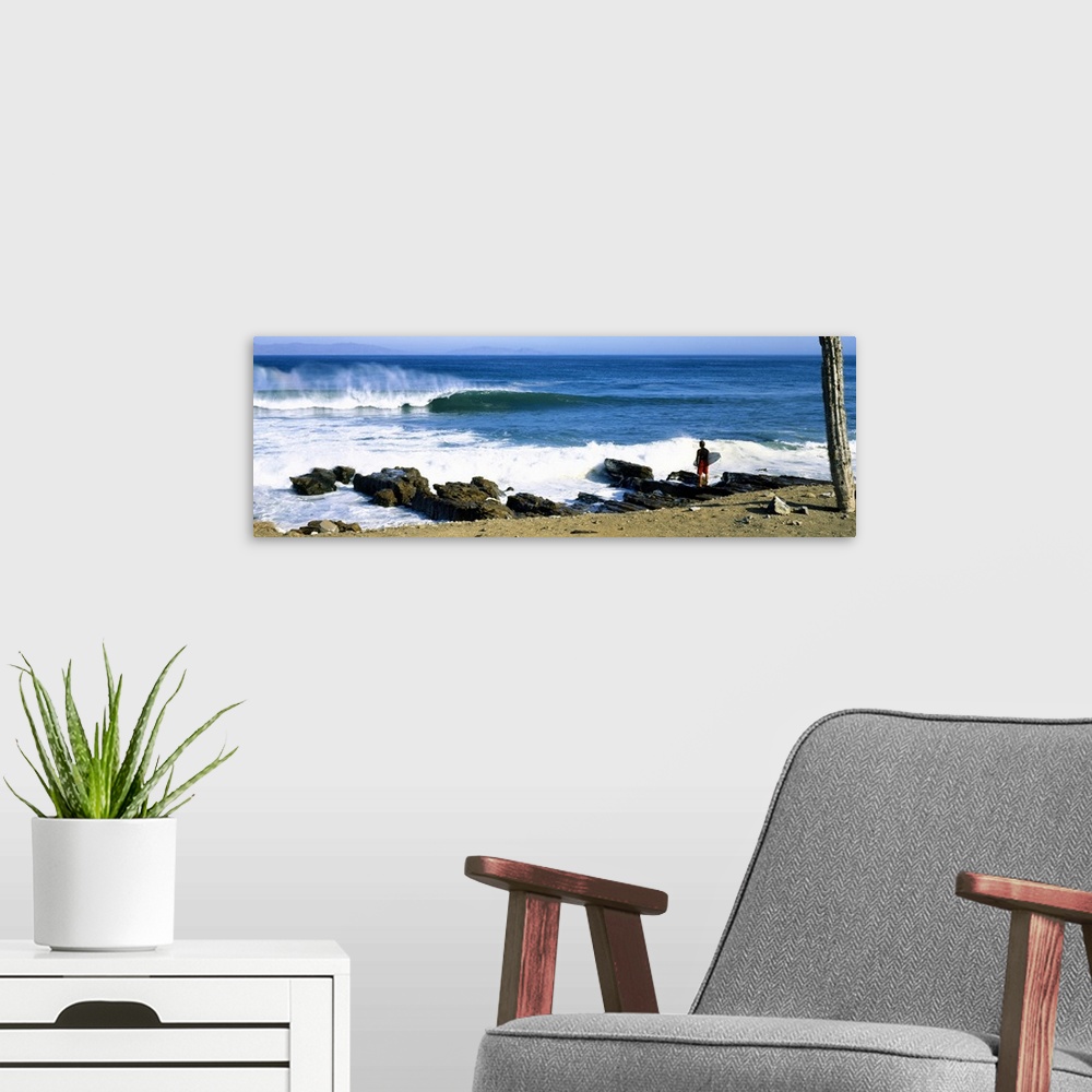 A modern room featuring A single figure stands amongst rocks watching waves break on the shore in this panoramic photograph.