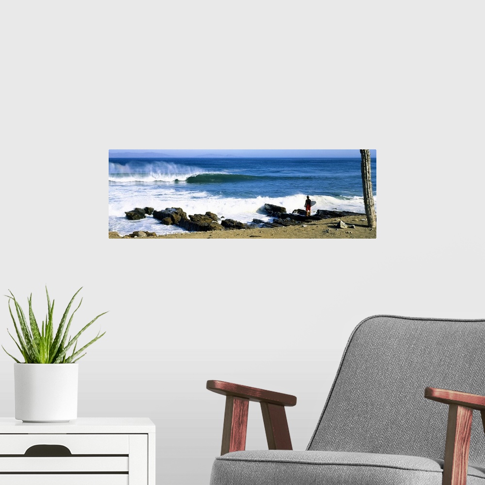 A modern room featuring A single figure stands amongst rocks watching waves break on the shore in this panoramic photograph.