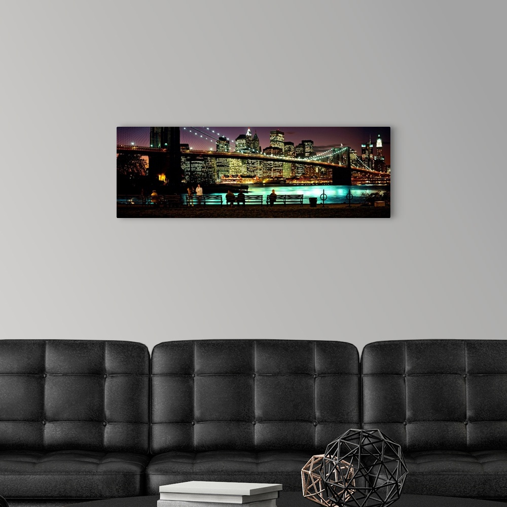 A modern room featuring Manhattan lights reflecting in the East River under the Brooklyn bridge while people sit on bench...