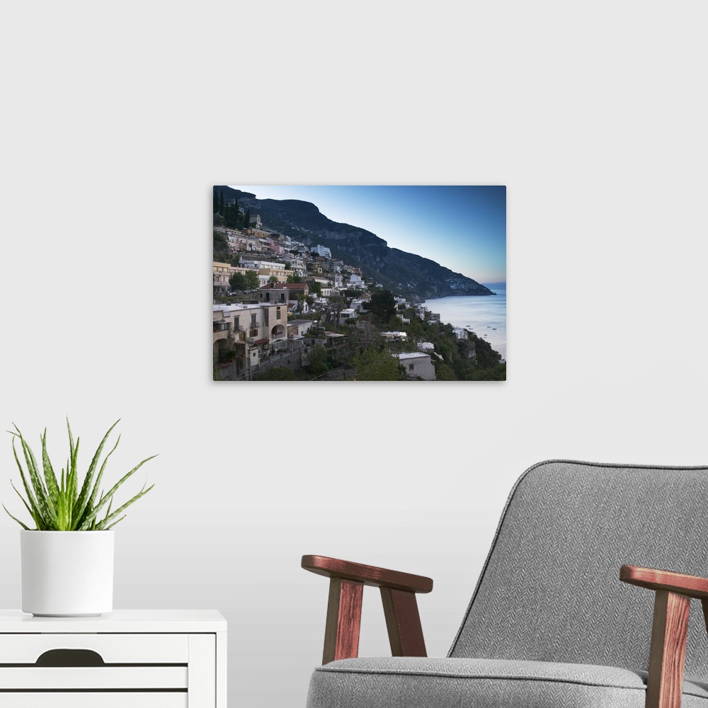 A modern room featuring Oversized artwork that is a picture of a town on a coast in Italy. A large hill is seen in the di...