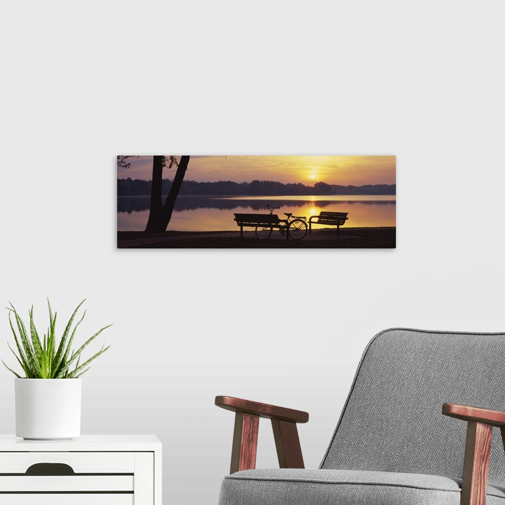 A modern room featuring Photograph of bike and park benches near waterfront with forest in the distance.  The sun is sett...