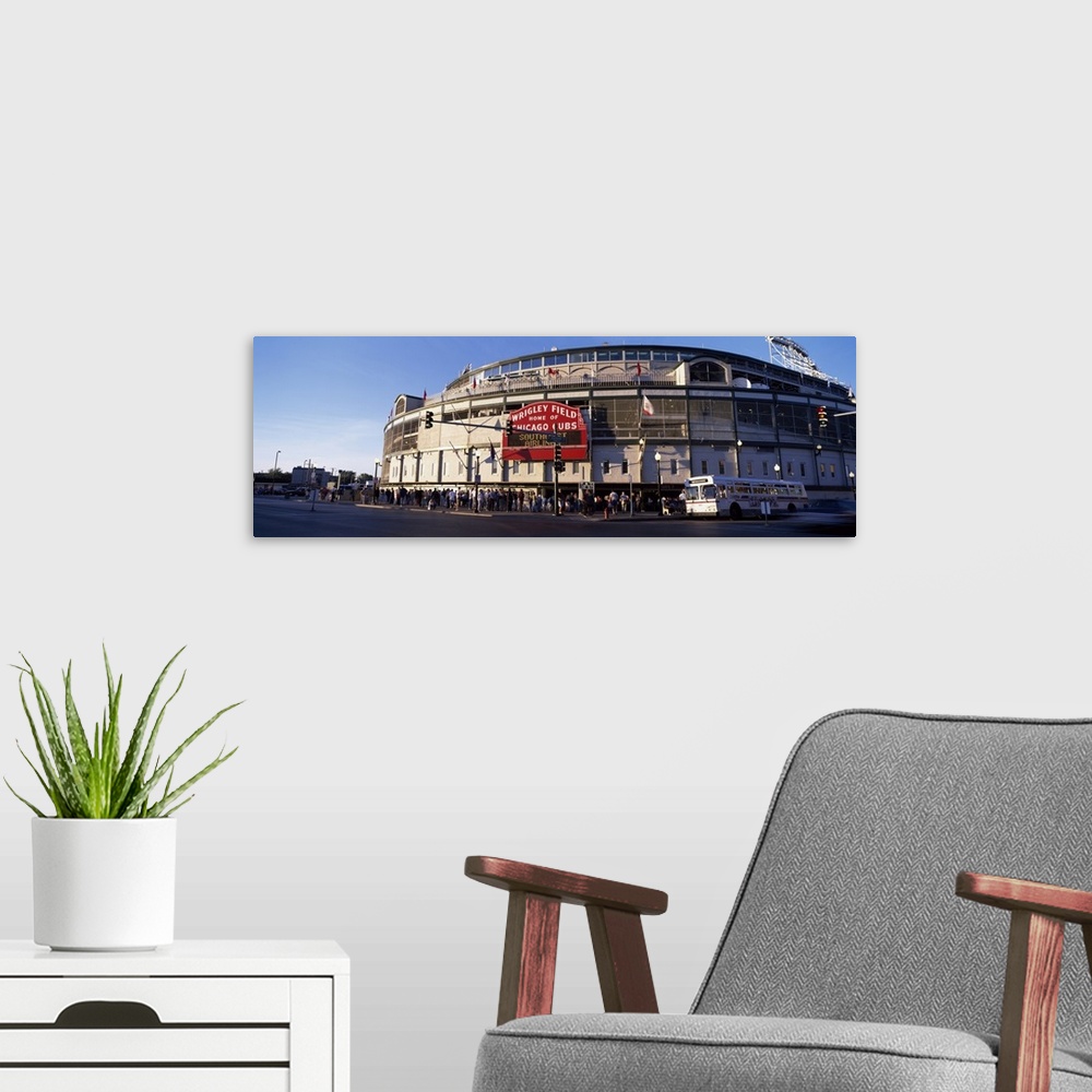A modern room featuring Wrigley Field is photographed in wide angle view with crowds standing in the front of the stadium...