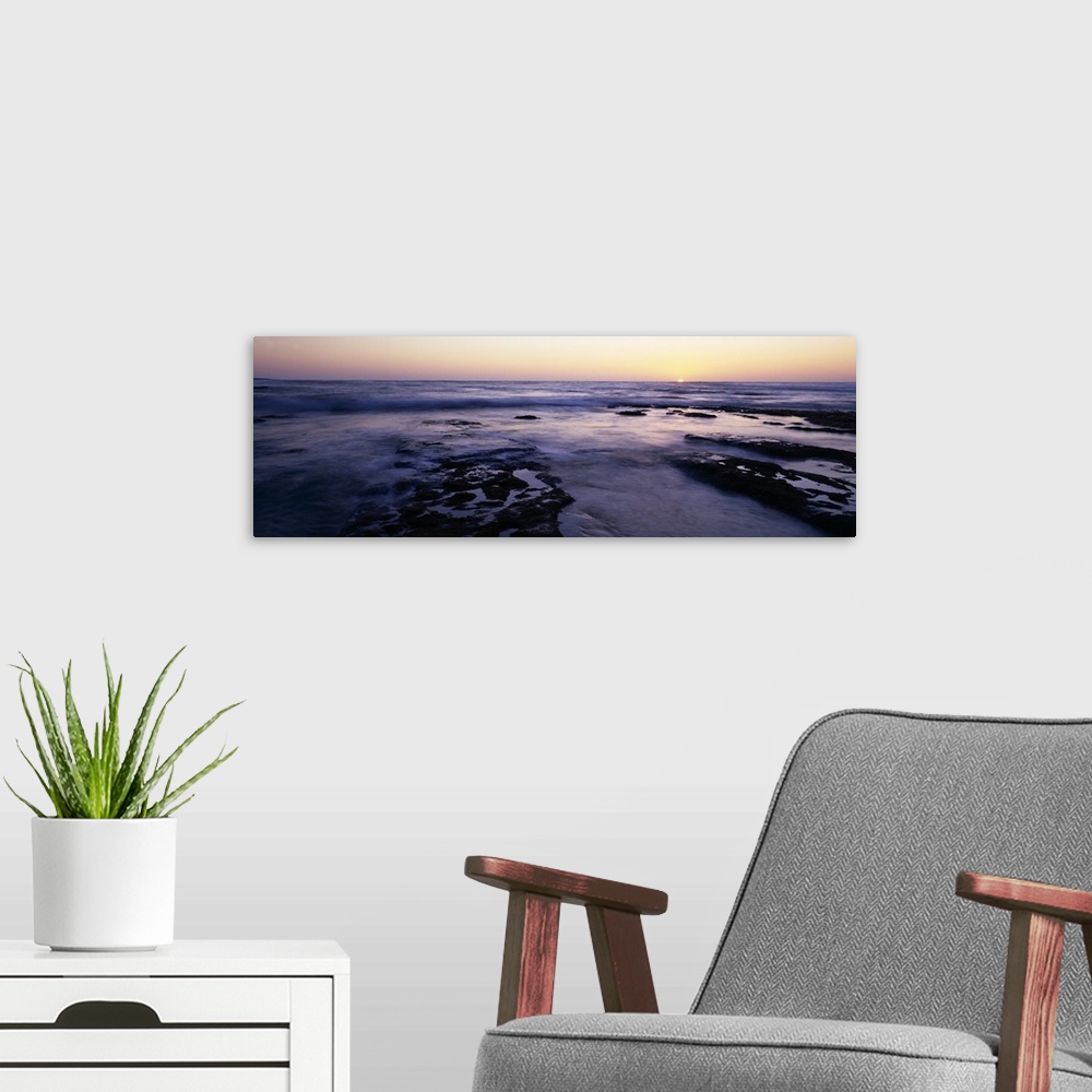 A modern room featuring Panoramic photograph taken of the ocean as the water flows over rocks and the sun is shown settin...