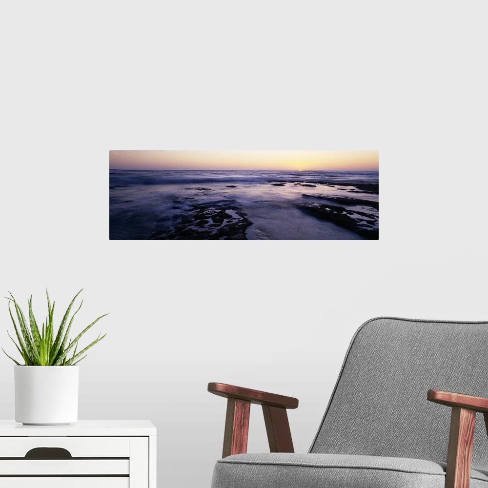 A modern room featuring Panoramic photograph taken of the ocean as the water flows over rocks and the sun is shown settin...