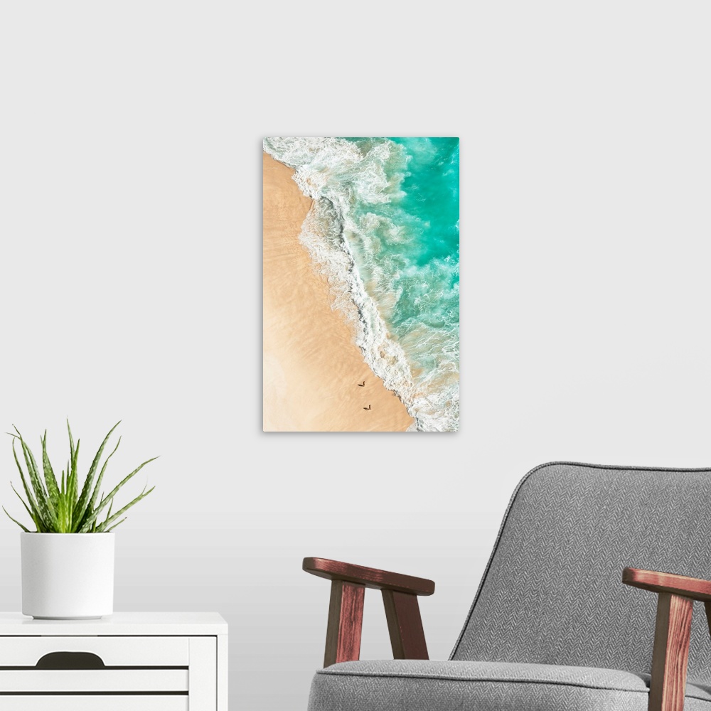 A modern room featuring A stunning overhead aerial photograph of bright turquoise waves crashing onto a sandy beach wth t...