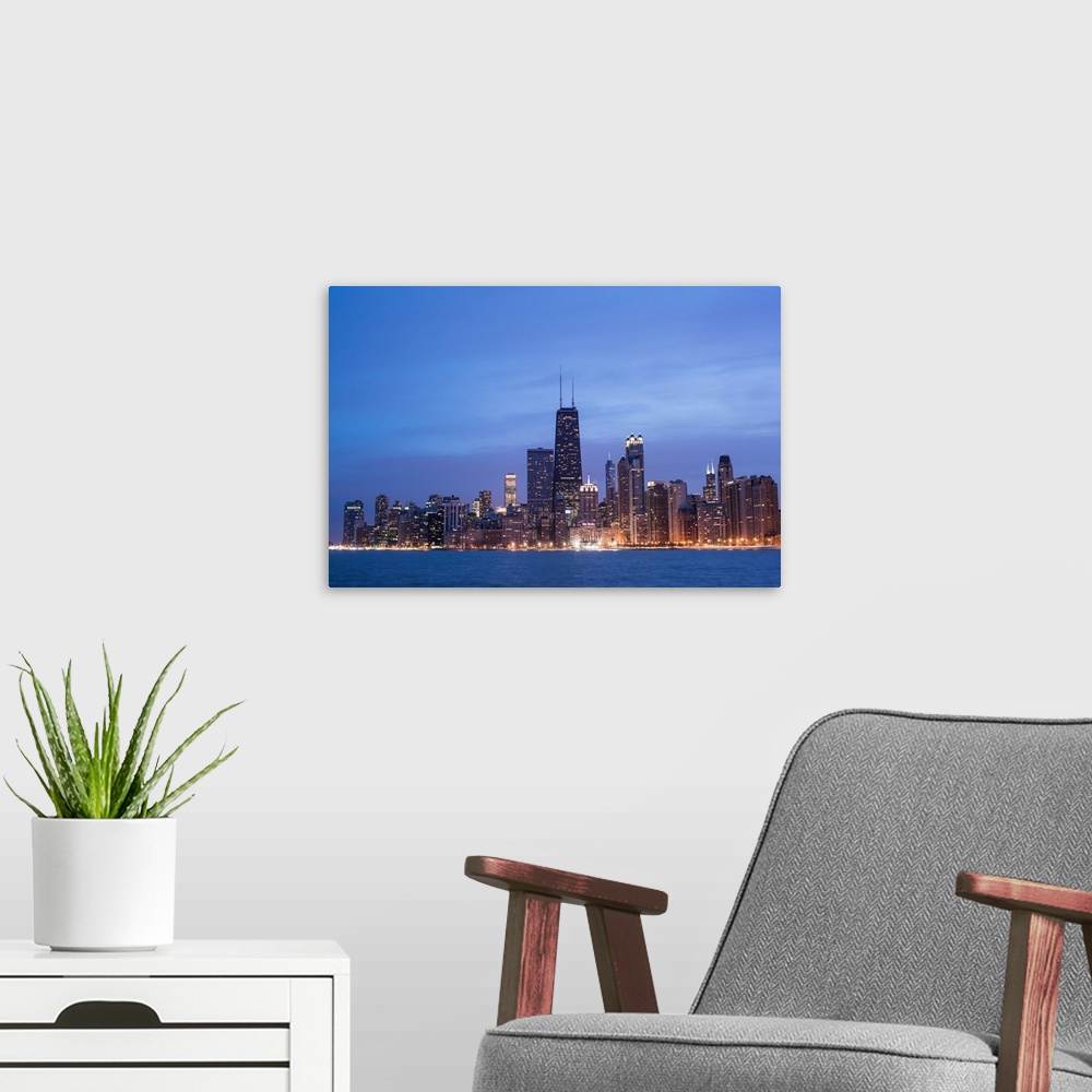 A modern room featuring The Chicago city skyline illuminated in the early evening, seen from across the water.