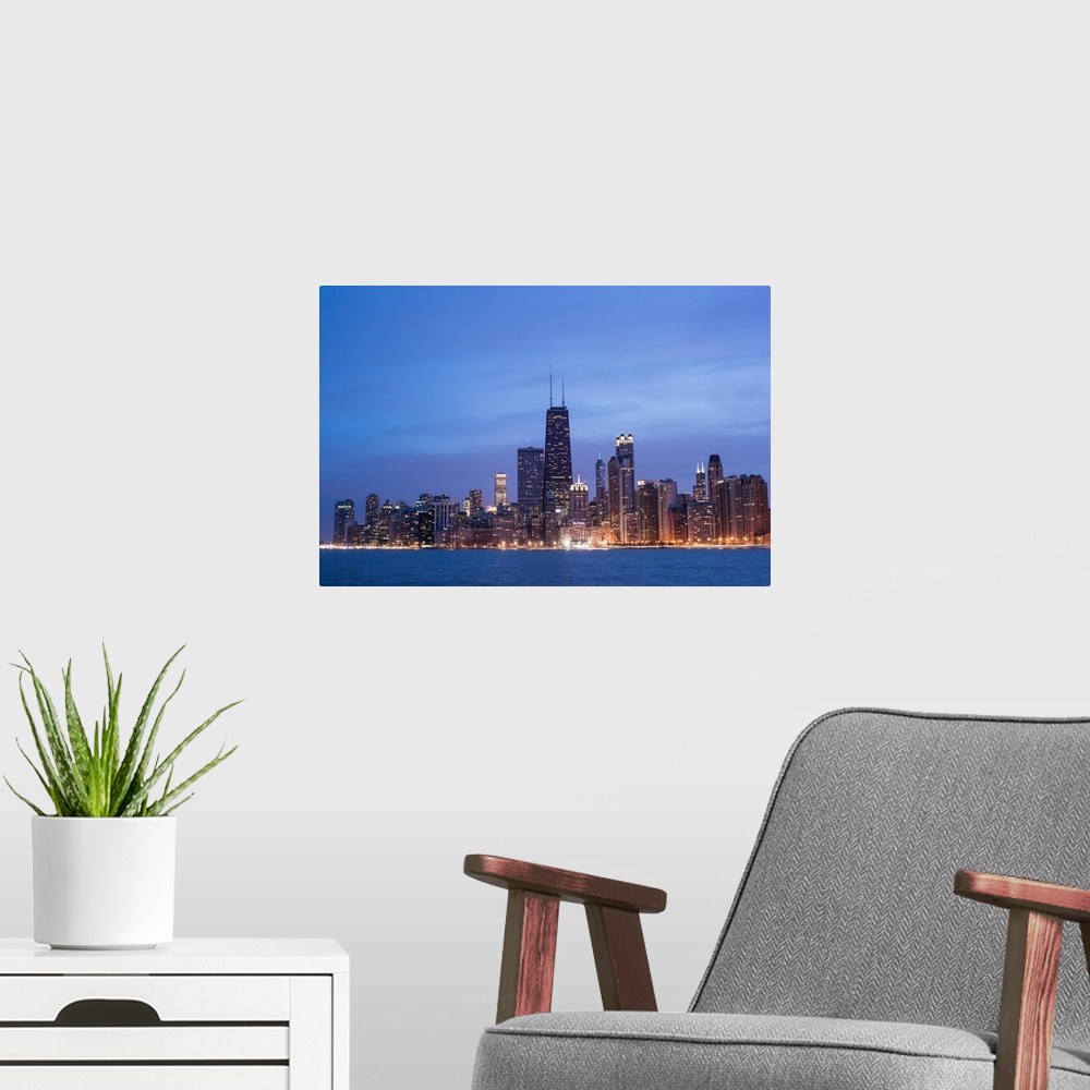 A modern room featuring The Chicago city skyline illuminated in the early evening, seen from across the water.