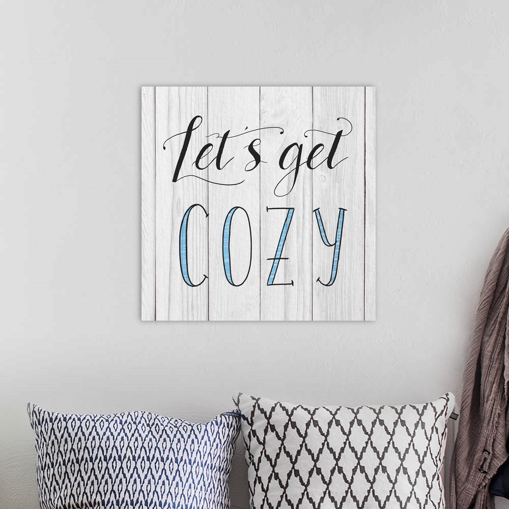 A bohemian room featuring "Let's Get Cozy" handwritten in black and blue on a white wood panel background.