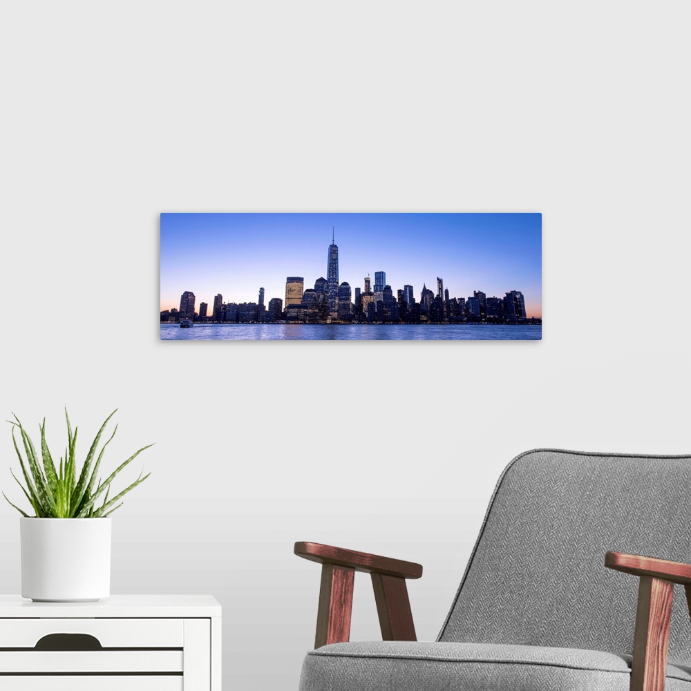 A modern room featuring Panoramic view of the New York City skyline with the One World Trade Center tower, at night.