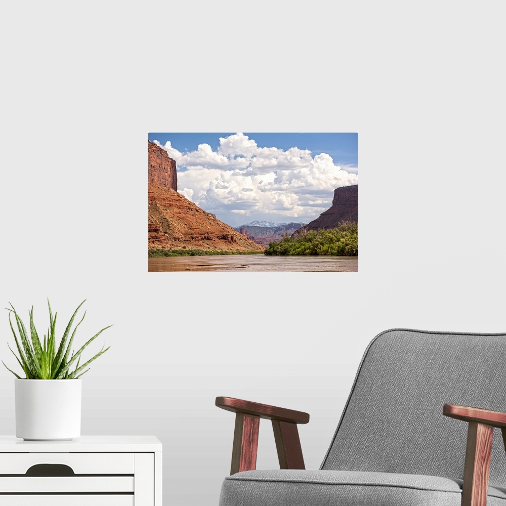 A modern room featuring Tall sandstone cliffs tower along the edge of the Colorado River, with clouds and blue skies over...