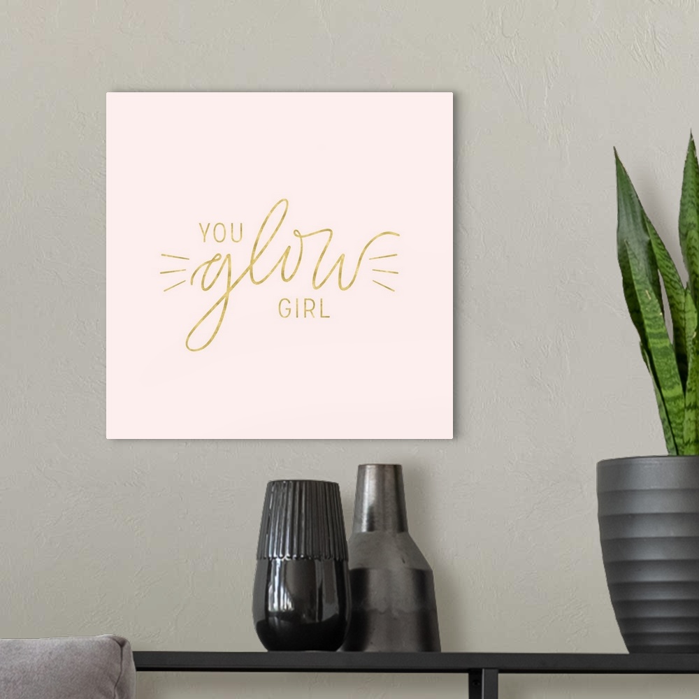 A modern room featuring "You Glow Girl" in metallic gold on a pale pink background.