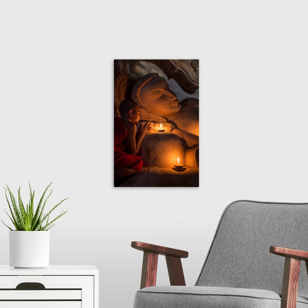 A modern room featuring Young Burmese monk praying by candlelight by large reclining Buddha.