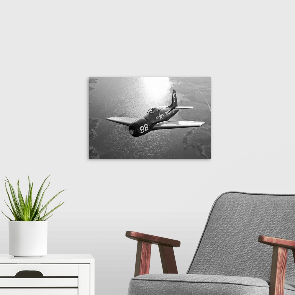 A modern room featuring A vintage military aircraft is photographed in black and white as it flies over a large body of w...