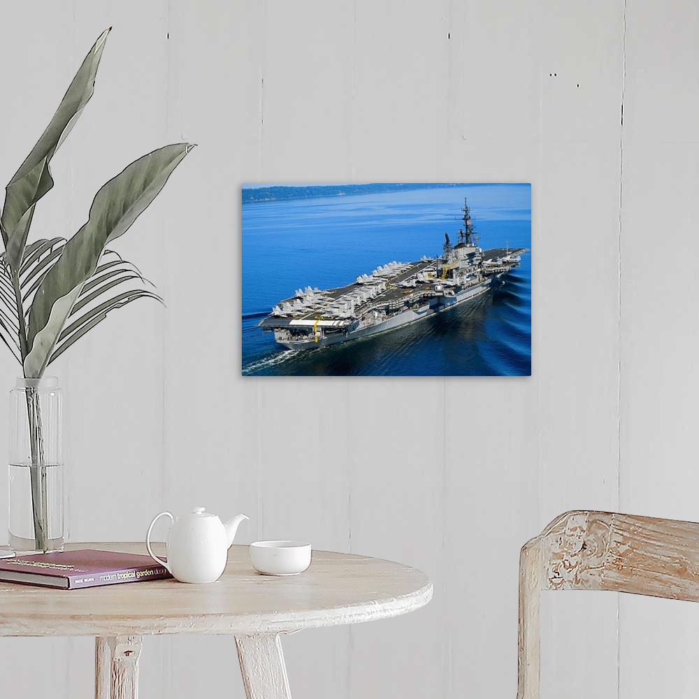 A farmhouse room featuring An aerial photograph of a naval warship not far off the coast its deck covered with fighter jets.