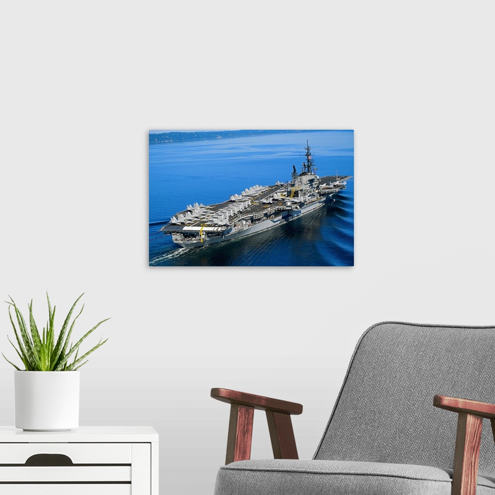 A modern room featuring An aerial photograph of a naval warship not far off the coast its deck covered with fighter jets.