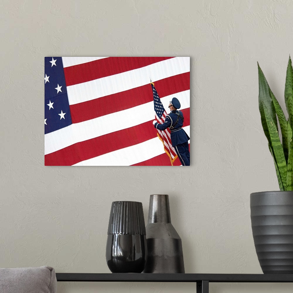 A modern room featuring A soldier stands in front of a massive American flag while holding a smaller flag during the chan...