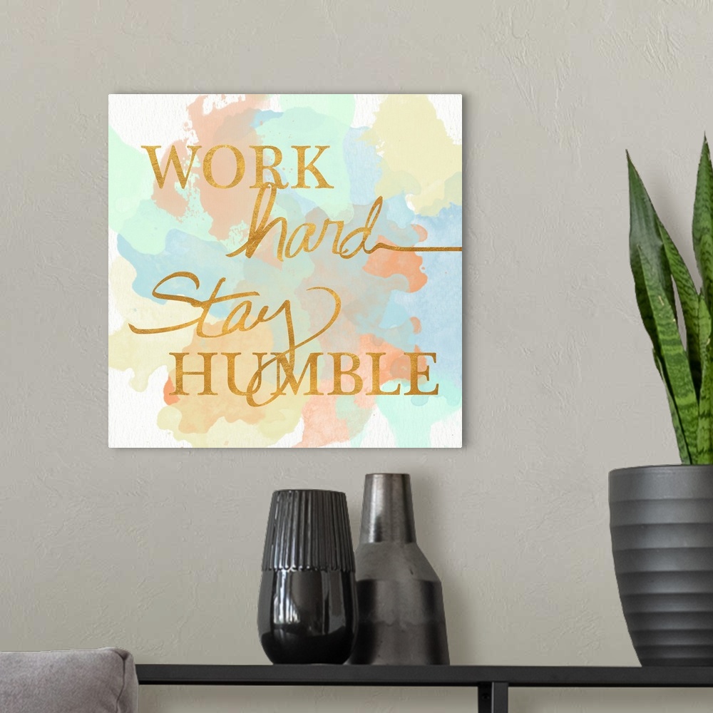 A modern room featuring "Work Hard Stay Humble"  written in a shiny gold font on a pastel colored watercolor background.