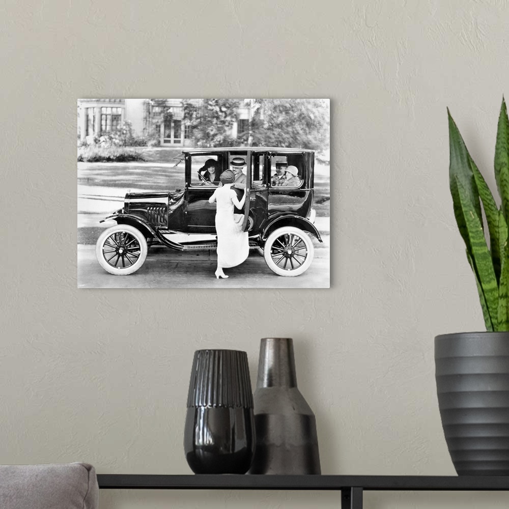 A modern room featuring A woman getting into a Ford Sedan with four other passengers. Photograph, c1923.