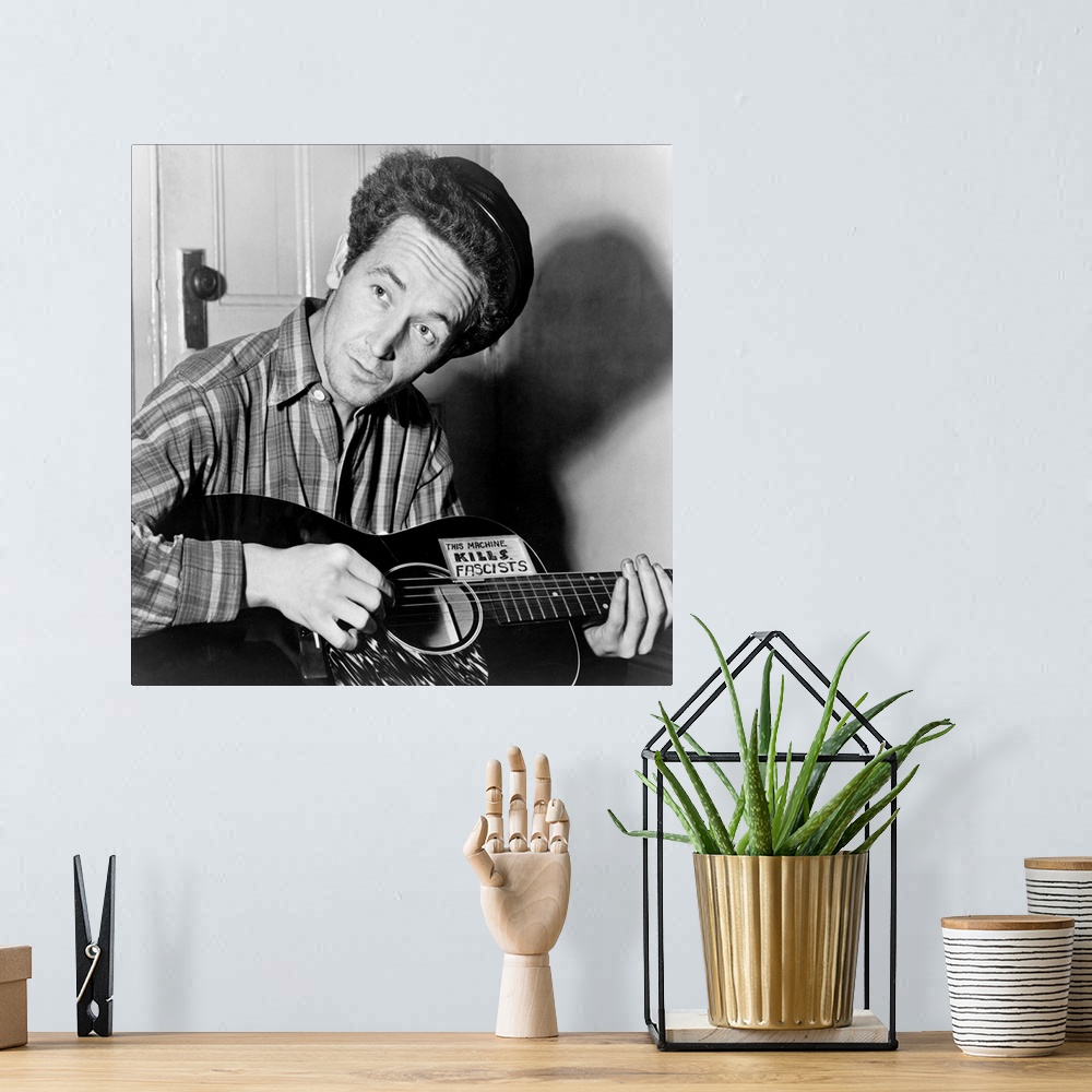 A bohemian room featuring American folk singer. Photographed playing a guitar that has a sticker attached reading: 'This Ma...