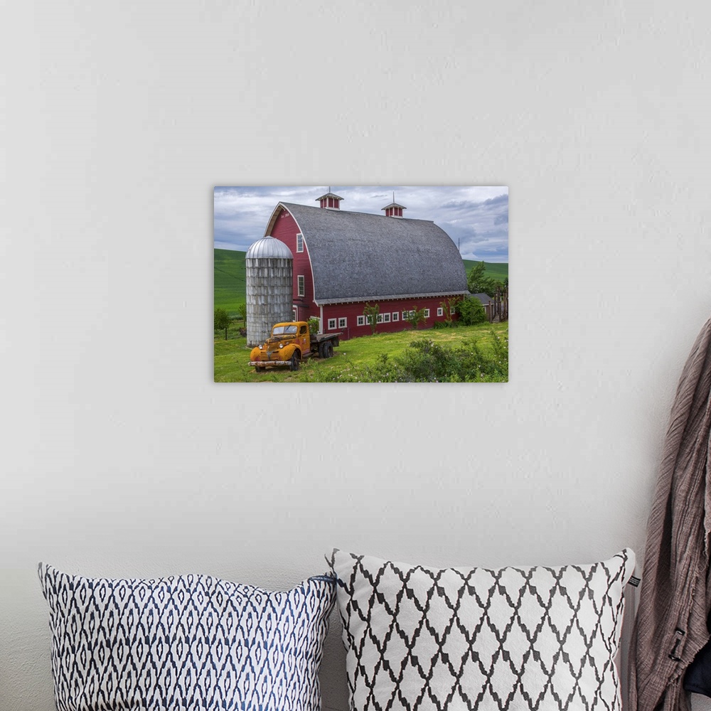 A bohemian room featuring A large red barn and an orange truck against the green landscape of Palouse, Washington.