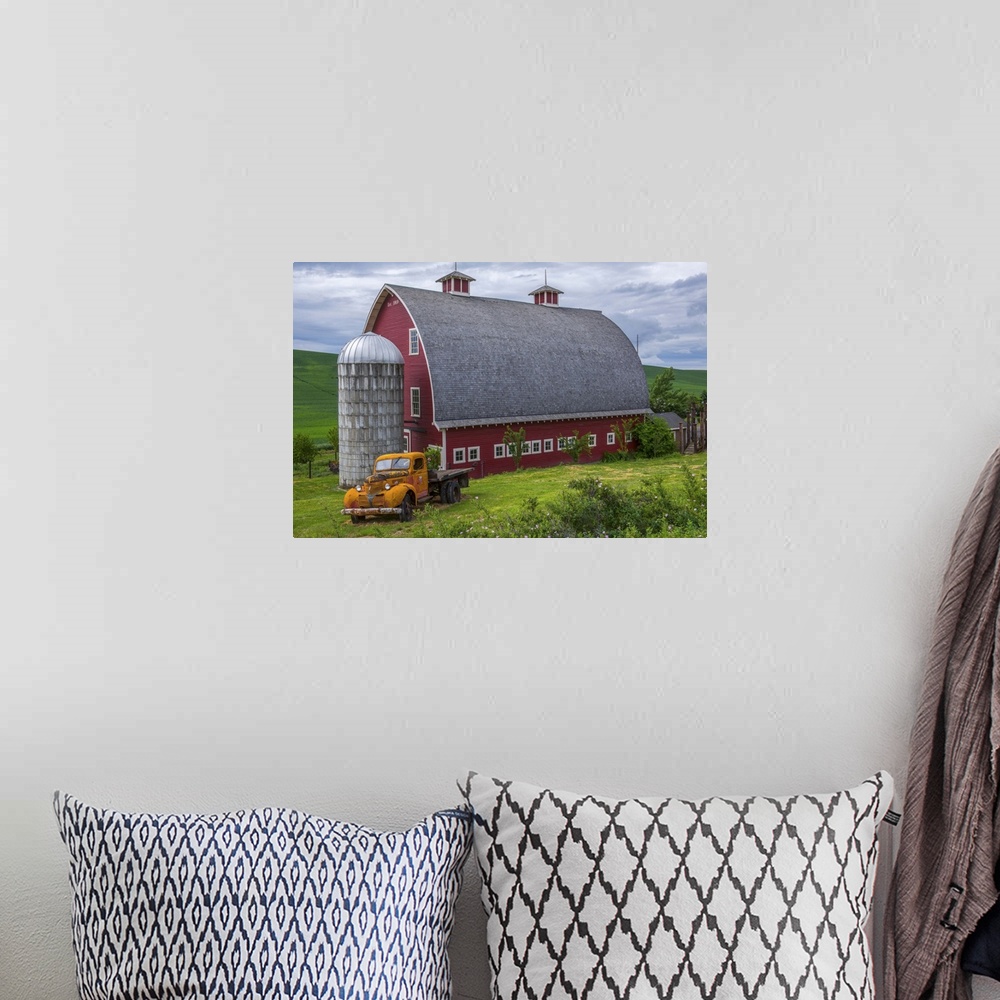 A bohemian room featuring A large red barn and an orange truck against the green landscape of Palouse, Washington.