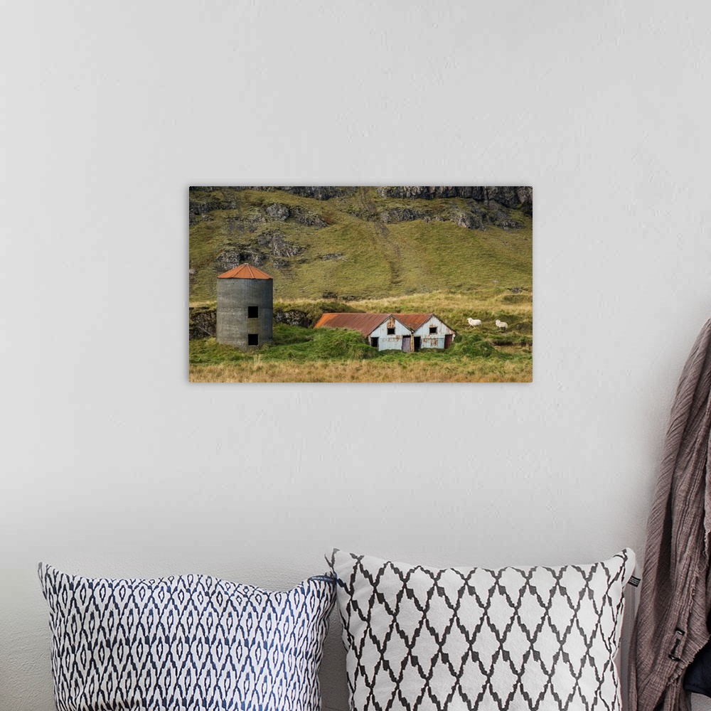 A bohemian room featuring Two sheep walking near rusted farmhouses and a silo at the bottom of a mountain in Iceland.