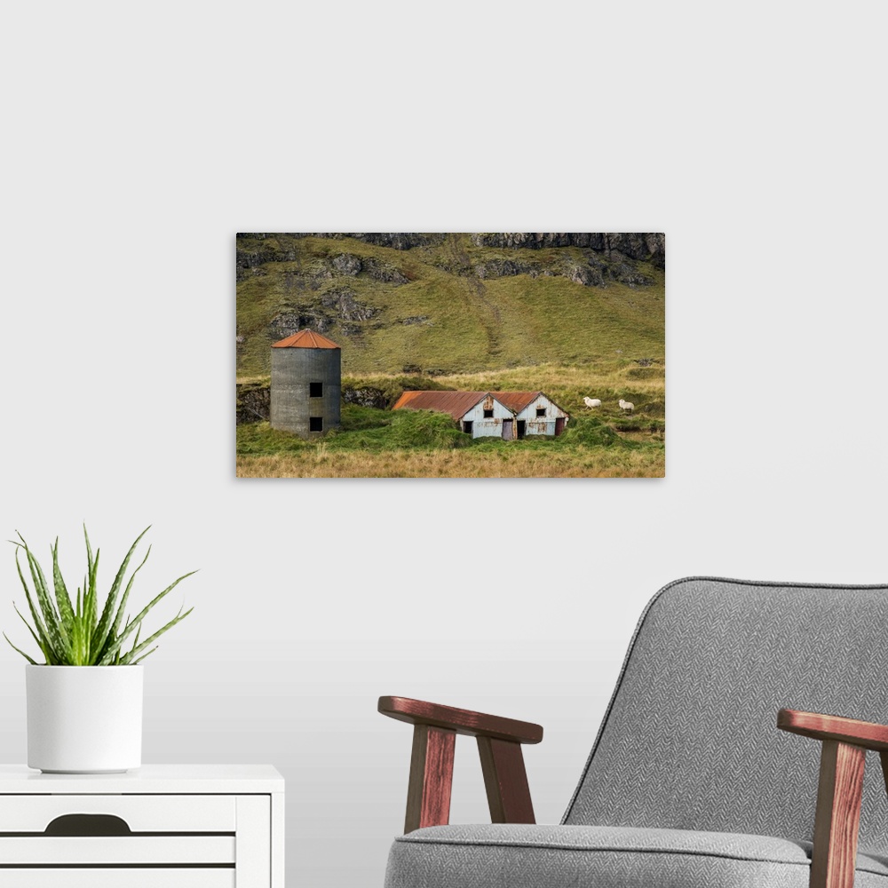 A modern room featuring Two sheep walking near rusted farmhouses and a silo at the bottom of a mountain in Iceland.