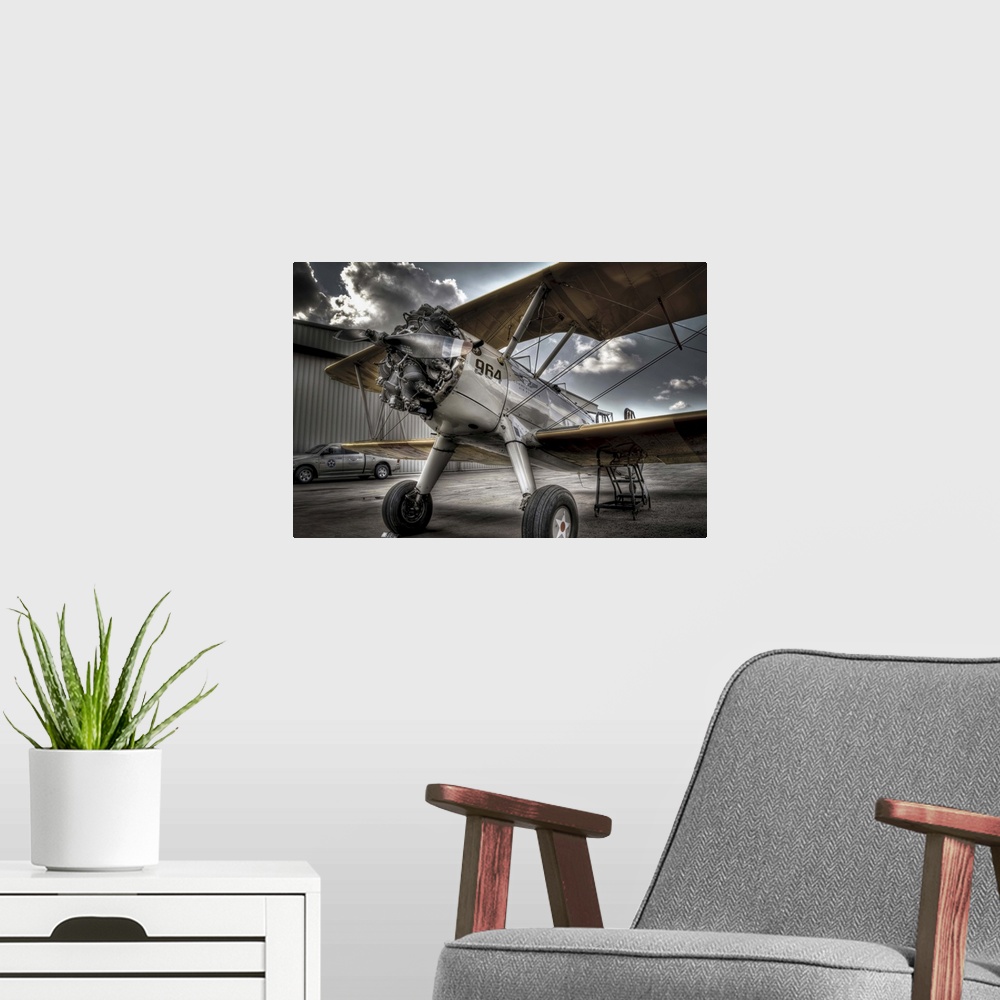 A modern room featuring A HDR photograph of a vintage air craft parked on the tarmac outside a hanger and a sky full of l...