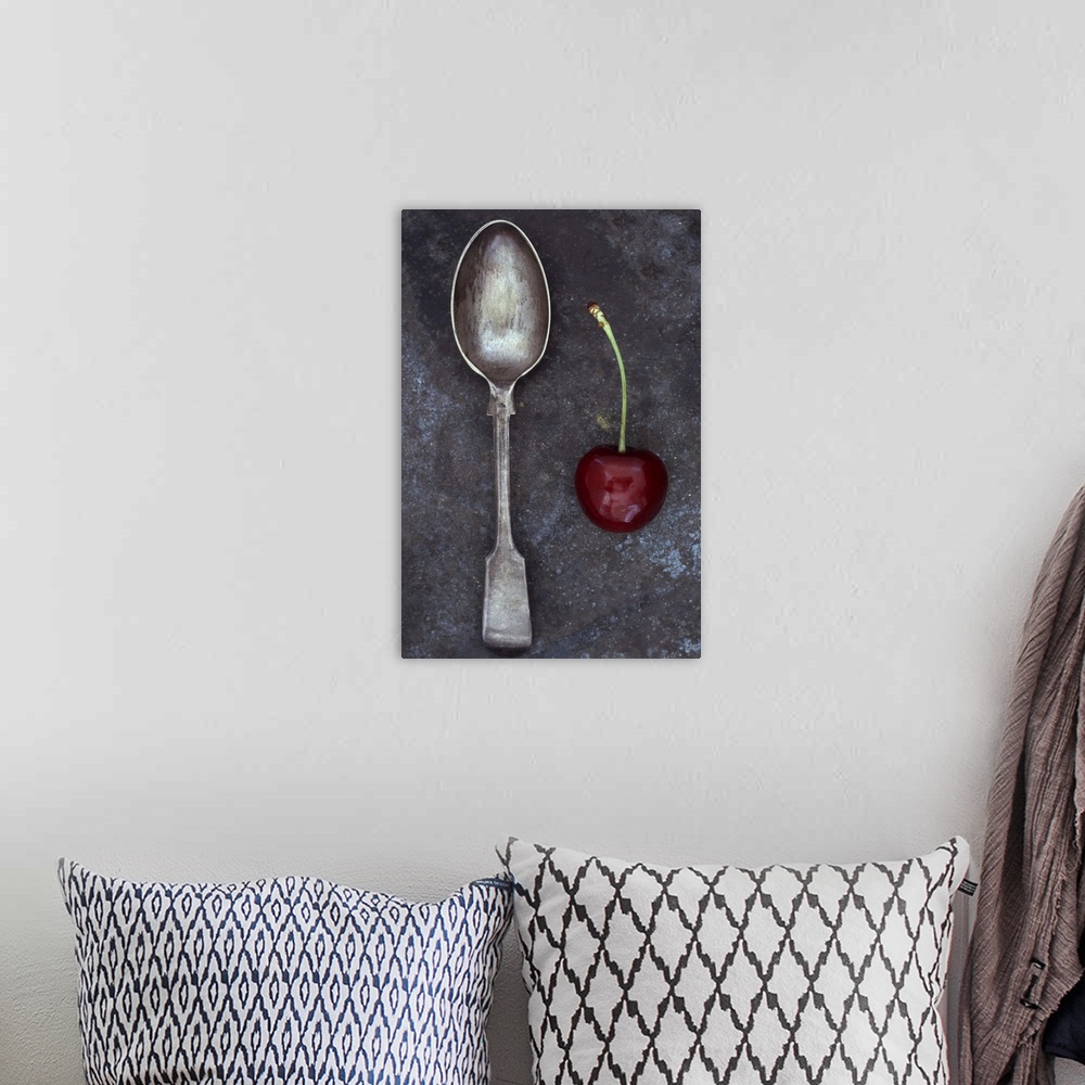 A bohemian room featuring Antique tarnished silver teaspoon lying next to single dark red cherry with stalk on tarnished metal