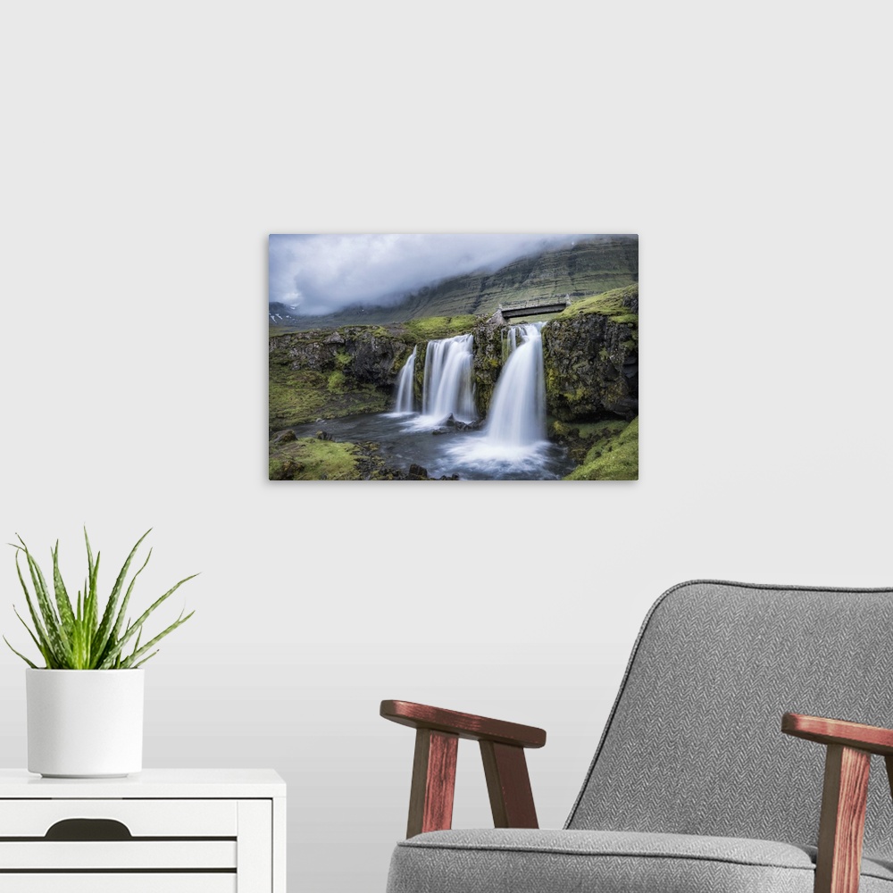 A modern room featuring Photograph of waterfalls from a river pouring into a small pool with a worn bridge and mountains ...