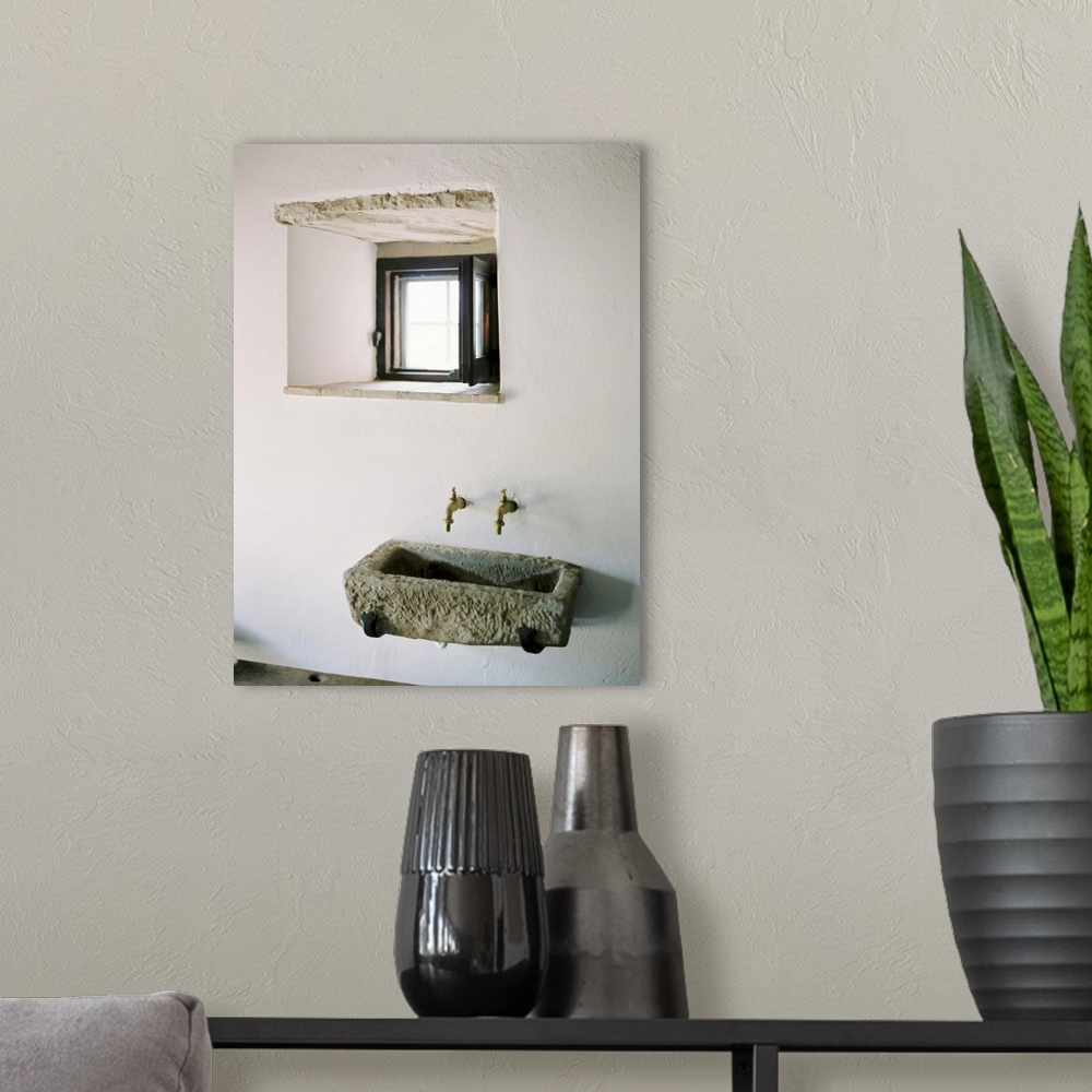 A modern room featuring A photograph of a rustic stone wall basin underneath a small window in a white stucco mediterrane...