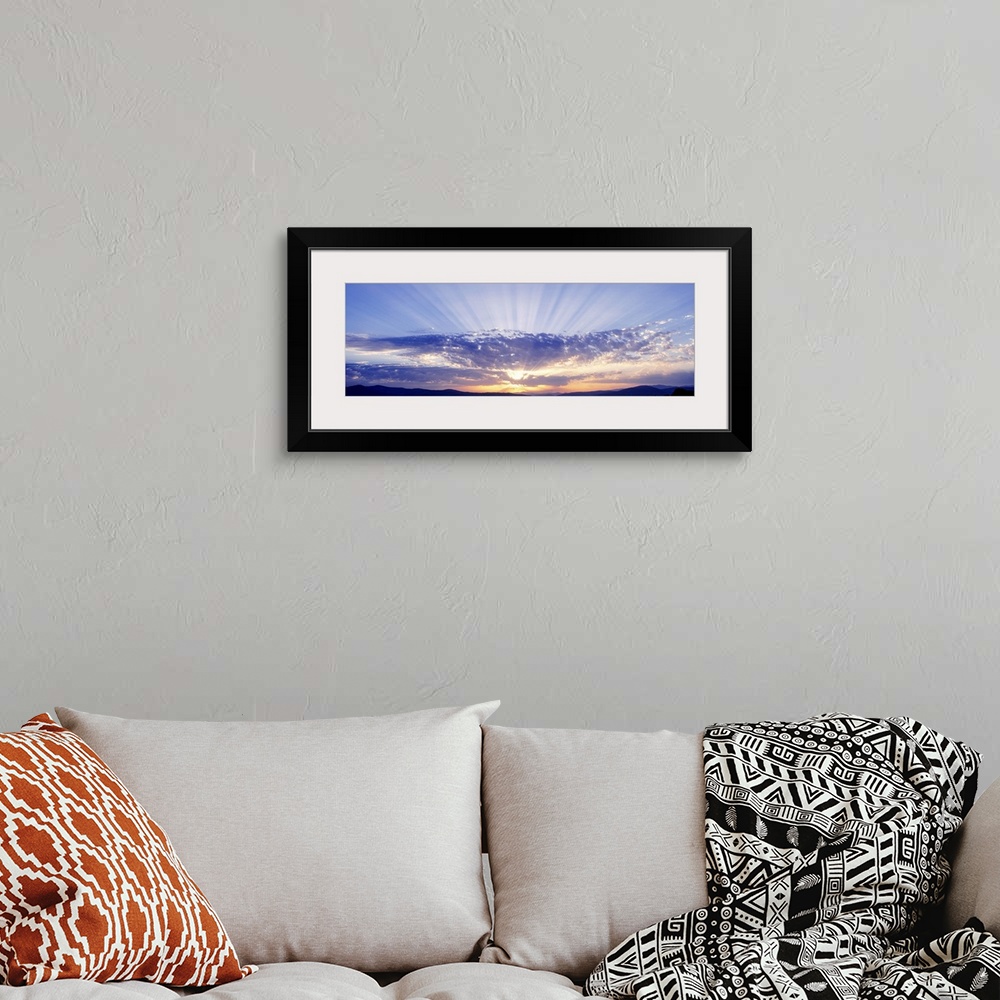 A bohemian room featuring This panoramic wall art captures as a photograph radiant sun beams radiate from behind clouds ove...