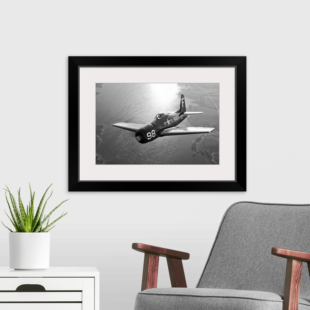 A modern room featuring A vintage military aircraft is photographed in black and white as it flies over a large body of w...
