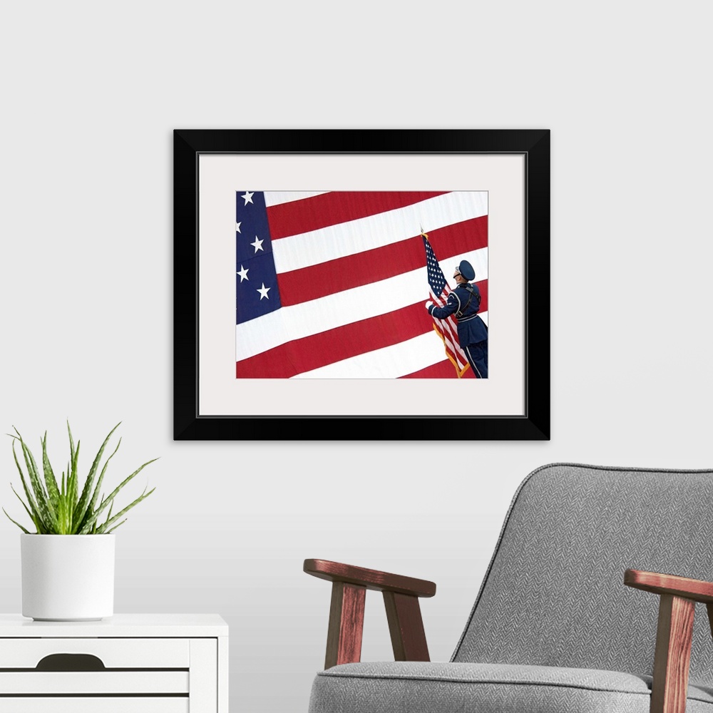 A modern room featuring A soldier stands in front of a massive American flag while holding a smaller flag during the chan...