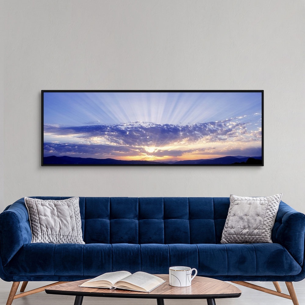 A modern room featuring This panoramic wall art captures as a photograph radiant sun beams radiate from behind clouds ove...