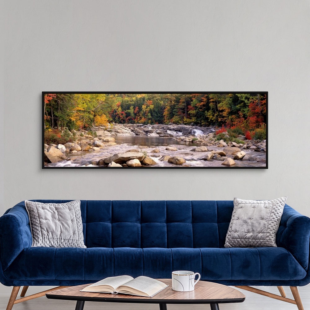 A modern room featuring A relaxed panoramic landscape of large boulders in a New England river lined with trees with autu...